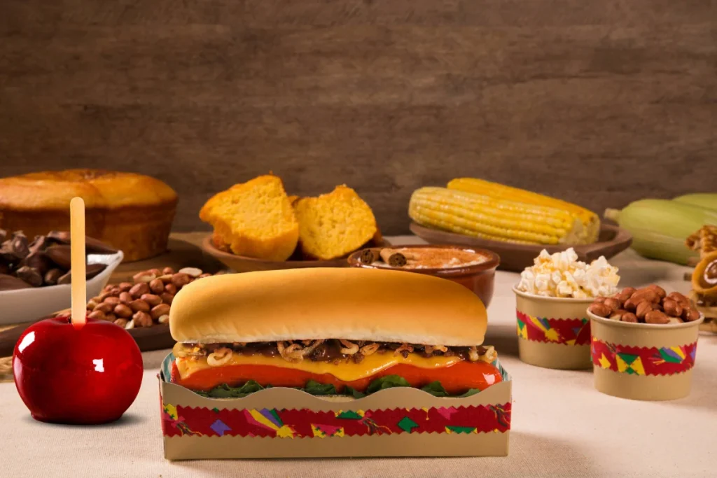  Registro de uma celebração de São João. A imagem captura a variedade de comidas típicas juninas, incluindo maçã do amor, pipoca doce, canjica, broa de fubá, milho verde cozido, entre outras receitas.