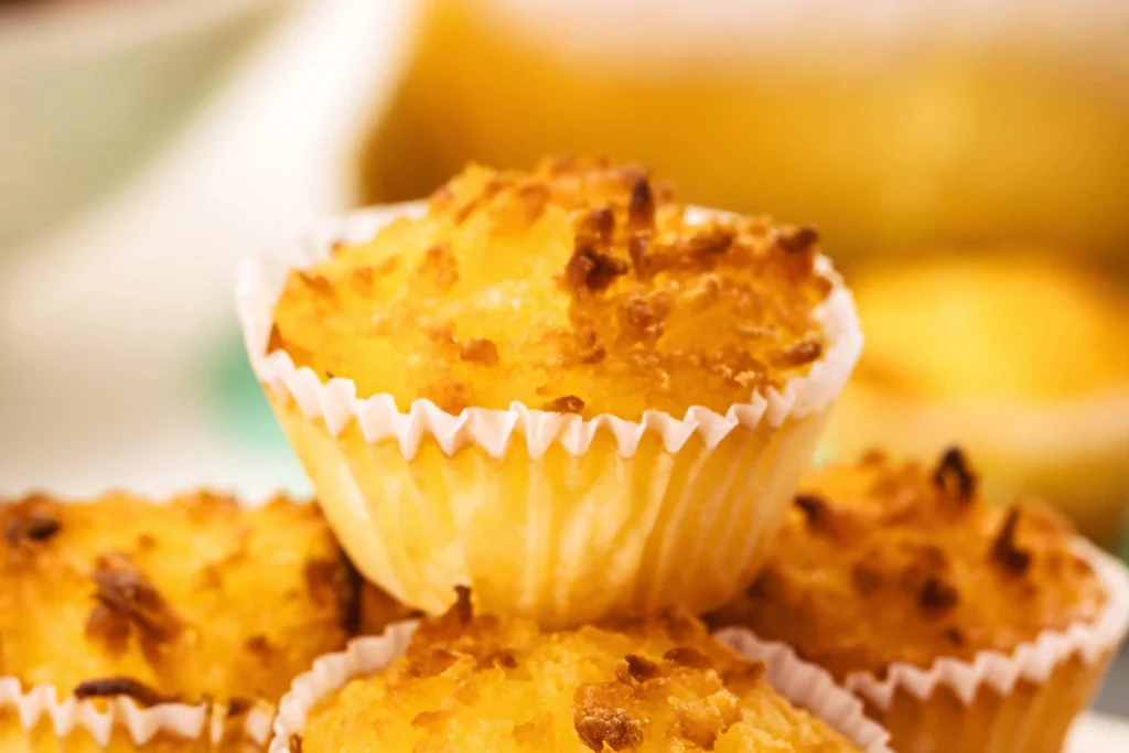Close-up de uma pilha de queijadinha de coco, ressaltando os fios de coco gratinados e crocantes no topo. O fundo está desfocado, mantendo a atenção na textura do doce.