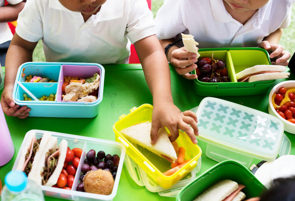 Crianças almoçando em uma escola primária, sentadas à mesa com lancheiras e pratos de comida.