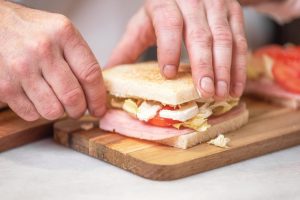 Close-up das mãos de uma pessoa preparando um sanduíche sobre uma tábua de cortar, com ingredientes frescos como alface e tomate visíveis.