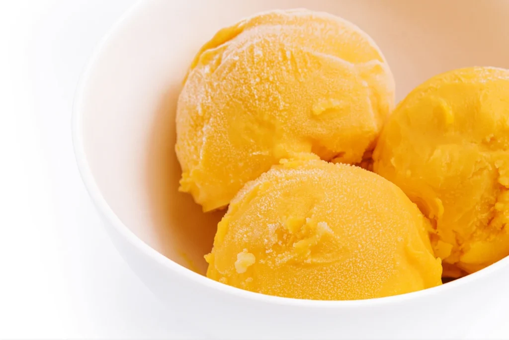 Como fazer sorvete de manga: 4 receitas fáceis de fazer em casa 2 Três porções arredondadas de sorvete de manga em uma tigela branca. É possível notar uma leve textura de congelamento na superfície, típica de sorvetes artesanais.