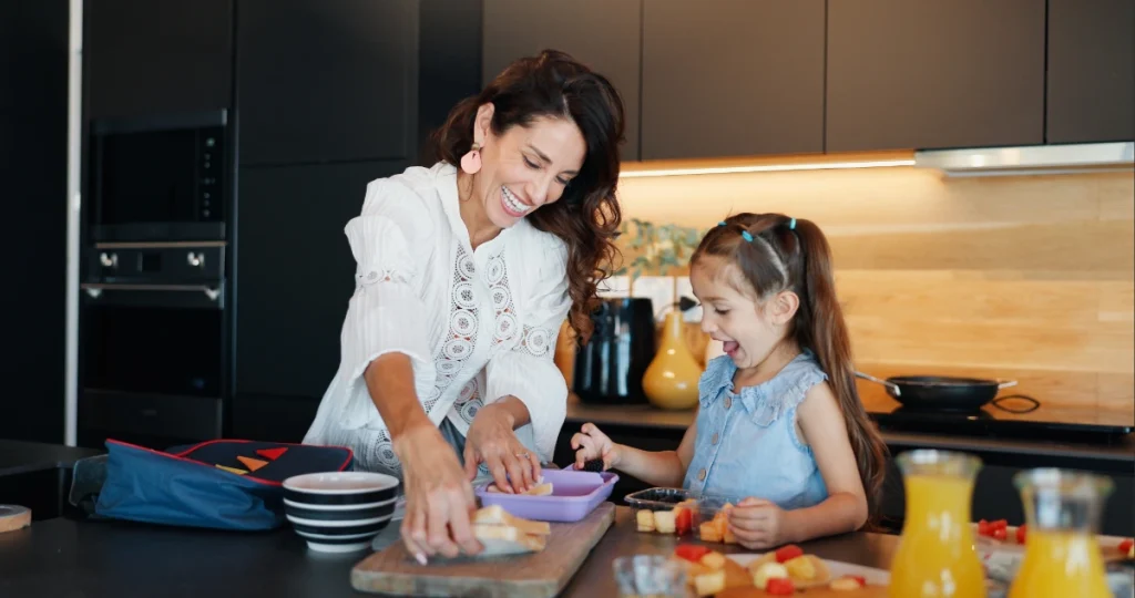 Mãe preparando lanche escolar para a filha em cima da bancada de uma cozinha.