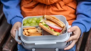 Criança segurando lancheira com lanche escolar, um sanduiche, frutas e biscoito