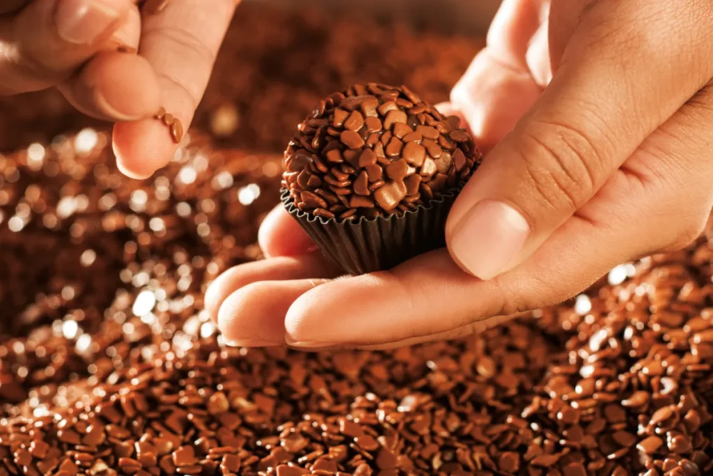 Uma pessoa segura com cuidado um brigadeiro em forminha preta. O fundo é composto por uma grande quantidade de confeitos de chocolate brilhantes, realçando a textura do doce.