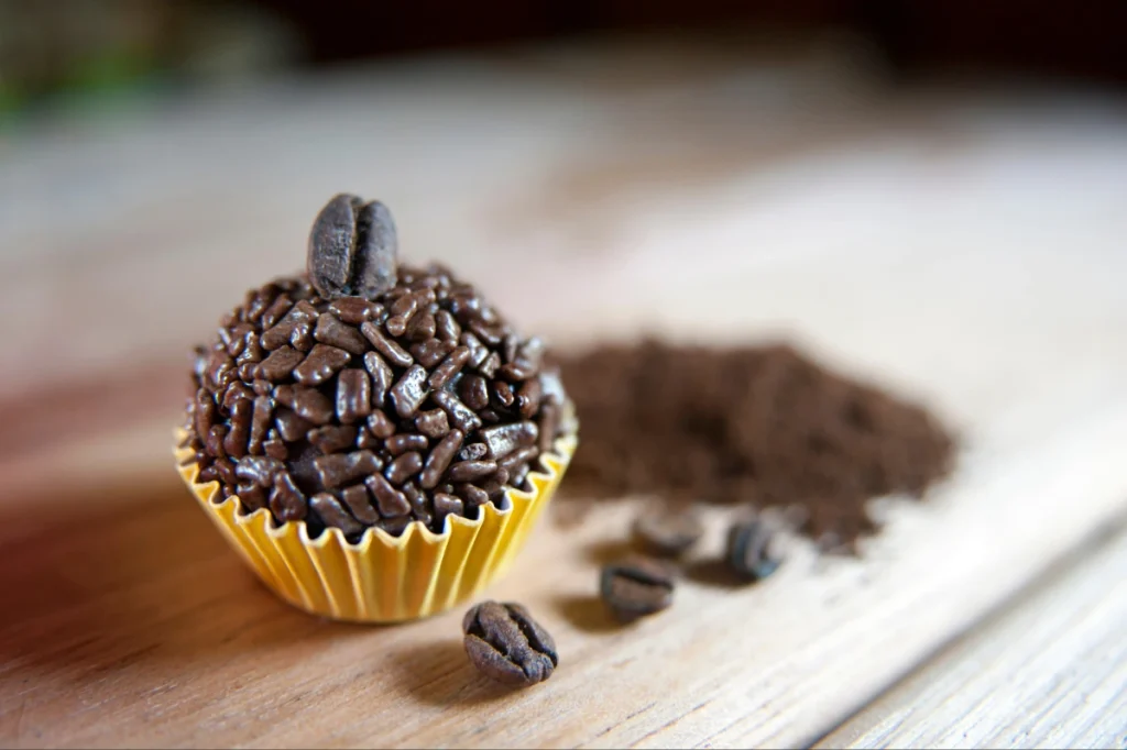 Brigadeiro de café sobre uma mesa de madeira com forminha amarela. Ao redor, grãos de café estão espalhados.