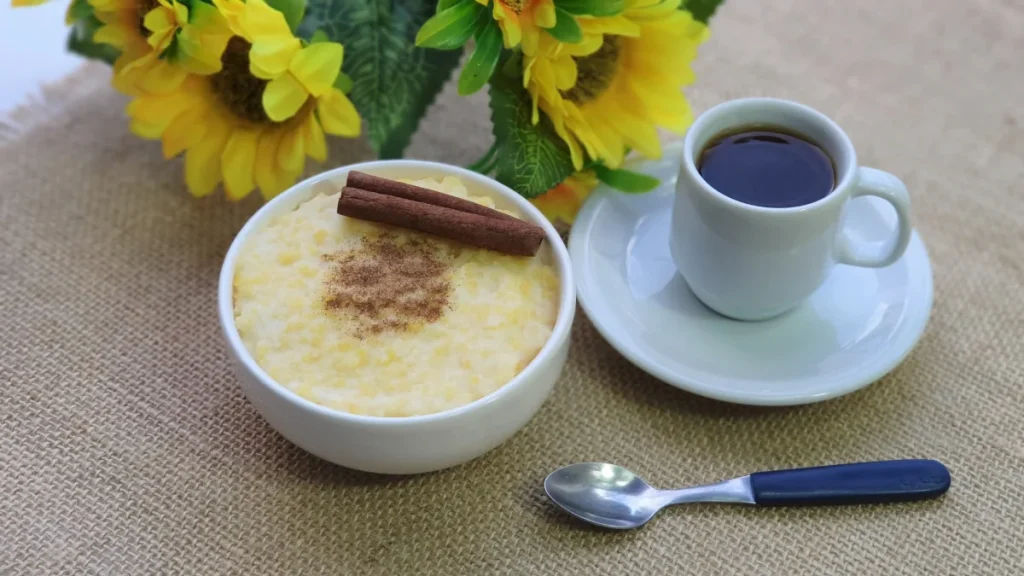 Canjica de milho doce com canela dentro de vasilha de louça branca ao lado de um xícara de café e flores artificiais amarelas.