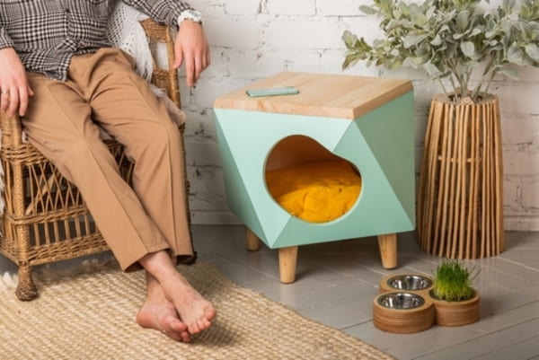 Cantinho pet com casinha verde menta em formato geométrico, entrada em coração e almofada amarela. Ao lado, dois potes de inox, grama para pets e planta decorativa. Ambiente interno com parede de tijolos brancos e tapete de fibras naturais.
