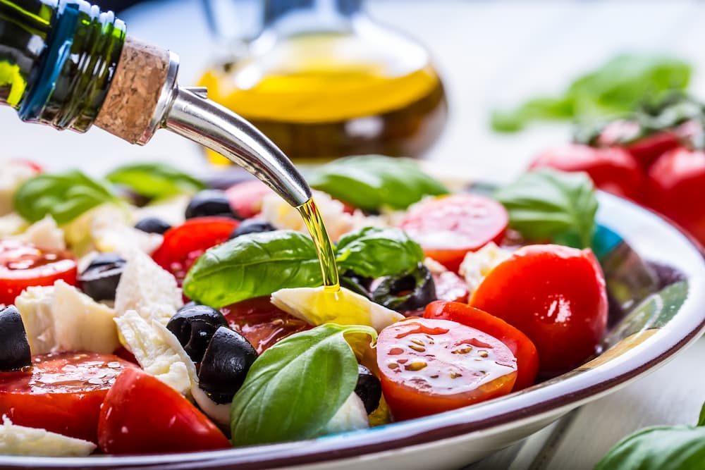 Salada colorida para uma alimentação saudável e saborosa! 1 Salada colorida fresca sendo preparada, com tomates-cereja, azeitonas pretas, folhas de manjericão e pedaços de queijo mussarela. Um fio de azeite está sendo derramado.
