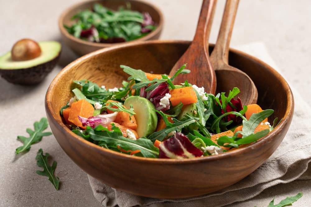 Salada colorida para uma alimentação saudável e saborosa! 2 Salada colorida servida em uma tigela de madeira, composta por folhas verdes como rúcula e radicchio, pedaços de batata-doce assada, fatias de abacate e queijo branco esfarelado.