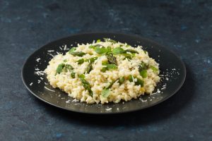 Prato de risoto cremoso com legumes, decorado com aspargos e ervas frescas, servido em prato escuro sobre fundo texturizado.