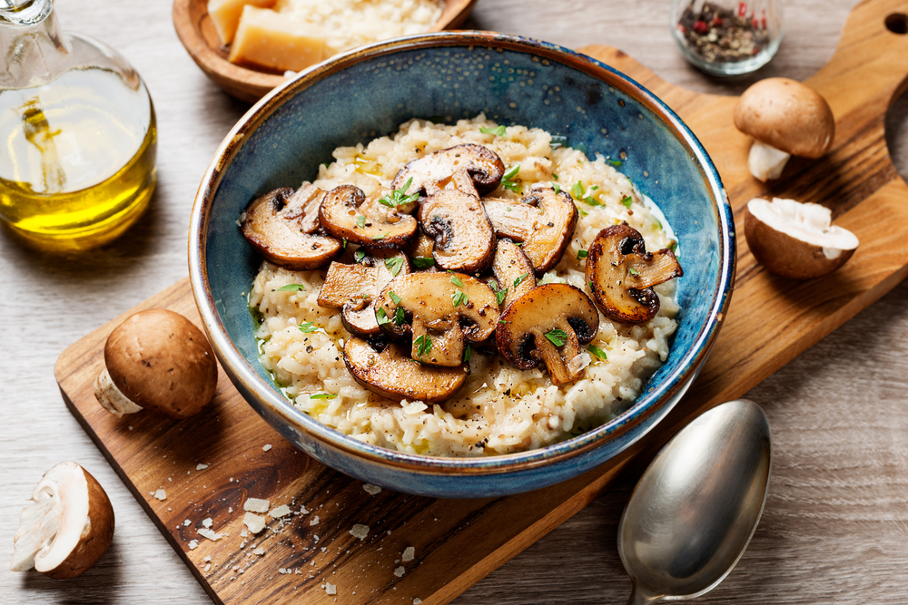 Risoto de legumes cremoso finalizado com cogumelos, servido em tigela sobre tábua de madeira, decorado com fatias de cogumelo salteado e ervas frescas.