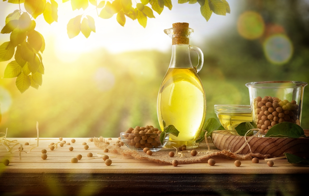 Frasco de óleo de soja ao lado de tigela com grãos de soja e recipiente com óleo, dispostos sobre superfície de madeira com folhas verdes e fundo natural desfocado.