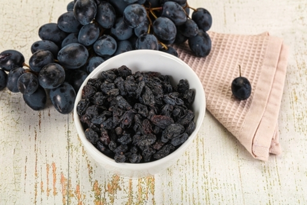 Saiba quais são os 11 alimentos proibidos para cães e gatos 2 Uvas frescas pretas e uvas-passas secas dispostas juntas sobre uma superfície branca, destacando o contraste entre a fruta fresca e a versão desidratada.