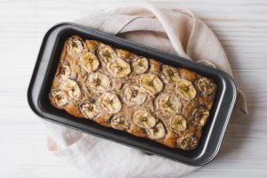 Bolo de banana caseiro recém-assado sobre uma tábua de madeira, com fatias cortadas que revelam sua textura macia e úmida, acompanhado de bananas maduras e nozes ao redor.