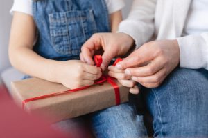 Menina sorridente entrega uma caixa de presente decorada com estrelas coloridas a uma professora sentada, que retribui com um sorriso caloroso. A cena ocorre em uma sala de aula infantil, com desenhos e bilhetes coloridos ao fundo, simbolizando carinho e gratidão pelo Dia dos Professores.