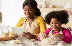 Mãe negra sorridente preparando uma receita com sua filha pequena na cozinha, ambas envolvidas na atividade culinária com alegria e carinho.