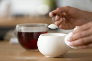 Pessoa adicionando adoçante a uma bebida quente, com uma colher retirando o produto de um recipiente branco, enquanto uma xícara de chá ou café aparece ao fundo sobre uma superfície de madeira.