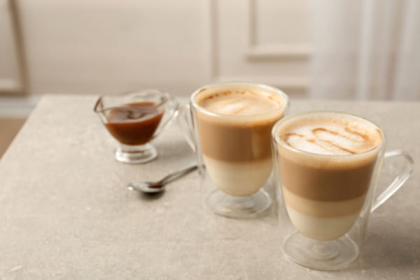Café cremoso com leite em pó: confira 3 receitas deliciosas 1 Duas xícaras de vidro com café cremoso, cobertas com uma camada espessa de espuma e calda de caramelo. As bebidas estão sobre uma superfície de madeira rústica, com um fundo desfocado que transmite uma atmosfera acolhedora de cafeteria.