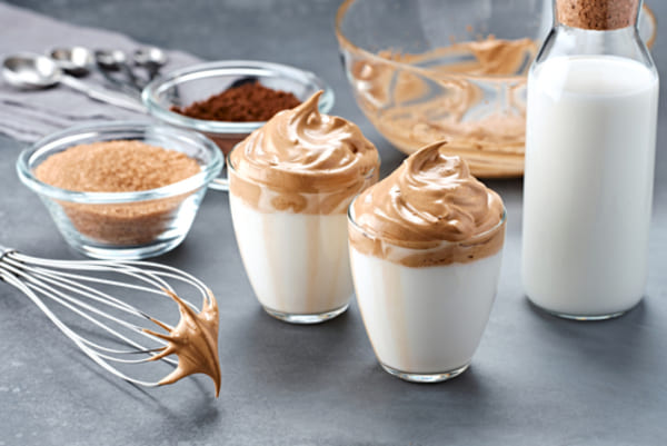 Café cremoso com leite em pó: confira 3 receitas deliciosas 3 Copo de vidro com café cremoso estilo dalgona, bebida coreana feita com café batido sobre leite, servido em uma superfície clara.