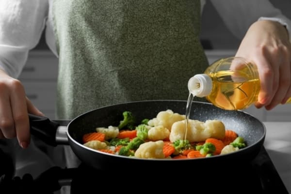 Mulher despejando óleo de cozinha de uma garrafa em uma panela com legumes cortados, sobre o fogão na cozinha. 