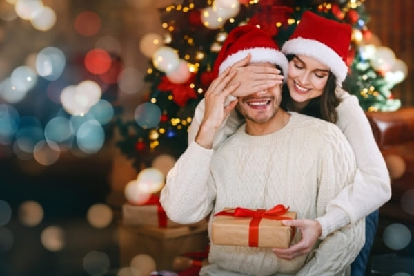 12 ideias de presentes de Natal para homens que nunca falham 13 Mulher jovem sorridente entregando um presente surpresa a um homem, em um ambiente acolhedor e iluminado, transmitindo carinho, afeto e o prazer de presentear.