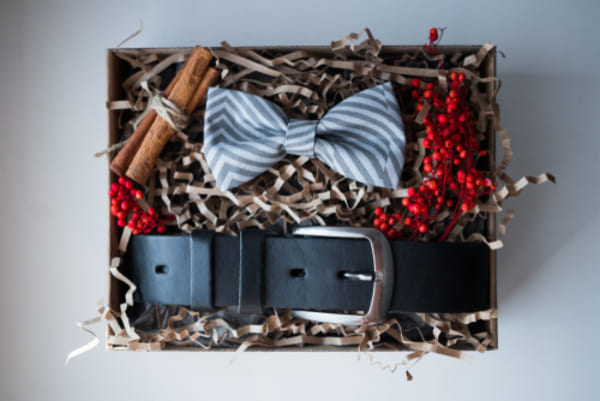 12 ideias de presentes de Natal para homens que nunca falham 4 Presentes elegantes com cinto, gravata borboleta e caixa de papel kraft sobre superfície de madeira, em composição sofisticada.