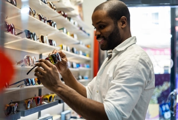 12 ideias de presentes de Natal para homens que nunca falham 9 Homem negro dentro de uma ótica escolhendo óculos de sol, examinando diferentes modelos com atenção.