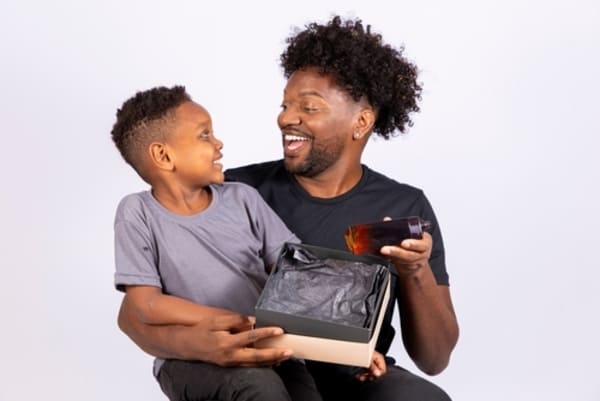 12 ideias de presentes de Natal para homens que nunca falham 7 Pai e filho negro comemorando o Dia dos Pais, com o menino entregando um presente embrulhado ao pai em um momento afetuoso.