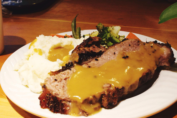 Vista superior de um bife servido em prato branco, acompanhado de molho de mostarda e purê de batatas, sobre uma mesa. 