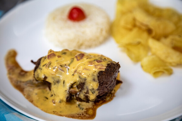 Vista superior de um bife grelhado ao molho de mostarda servido em prato branco, acompanhado de batatas chips e arroz. 
