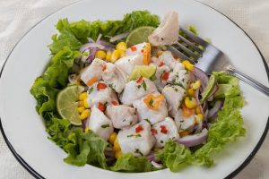 Prato típico peruano de ceviche de peixe, servido em uma tigela branca, com pedaços de peixe cru marinados em suco de limão, cebola roxa, pimenta e ervas frescas, acompanhado de milho e batata-doce.
