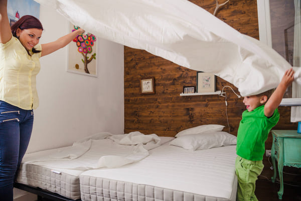 Como manter a casa limpa e organizada? Produtos e truques para um lar aconchegante 1 Mulher e criança estendendo o lençol sobre a cama, organizando o quarto para deixá-lo organizado e aconchegante.