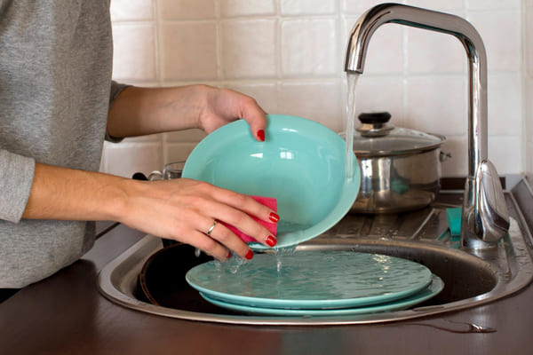 Como manter a casa limpa e organizada? Produtos e truques para um lar aconchegante 2 Mulher lavando pratos na pia da cozinha em casa após a refeição, representando rotina doméstica e cuidado com a limpeza.