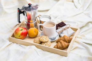 Café da manhã servido sobre lençóis bagunçados, com uma bandeja contendo xícara de café, suco, frutas e croissants, criando um clima aconchegante e casual.