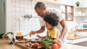 Pai e filho na cozinha ao lado de ingredientes e um notebook, preparando um Almoço de Dia das Mães simples.