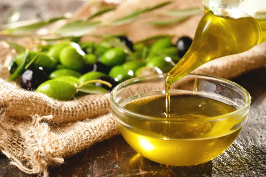 Azeite de oliva extra virgem sendo despejado em uma tigela de vidro com azeitonas ao fundo.
