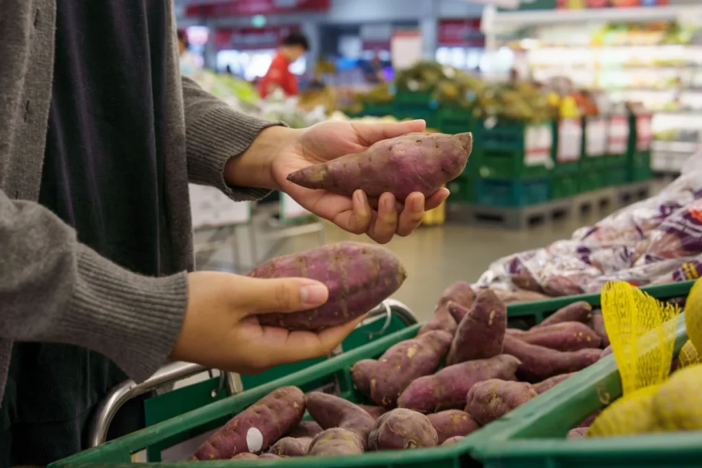  Mãos de uma pessoa escolhendo e segurando duas batatas doces de casca roxa em uma seção de hortifruti, representando o cuidado na seleção dos ingredientes.