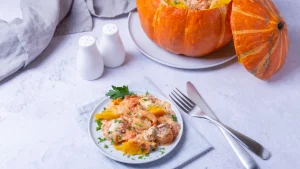 Close-up de um prato individual de camarão na moranga com molho alaranjado e pedaços de polpa de abóbora, decorado com salsa, um prato típico do outono no Brasil. Ao lado, talheres de metal sobre uma mesa clara.