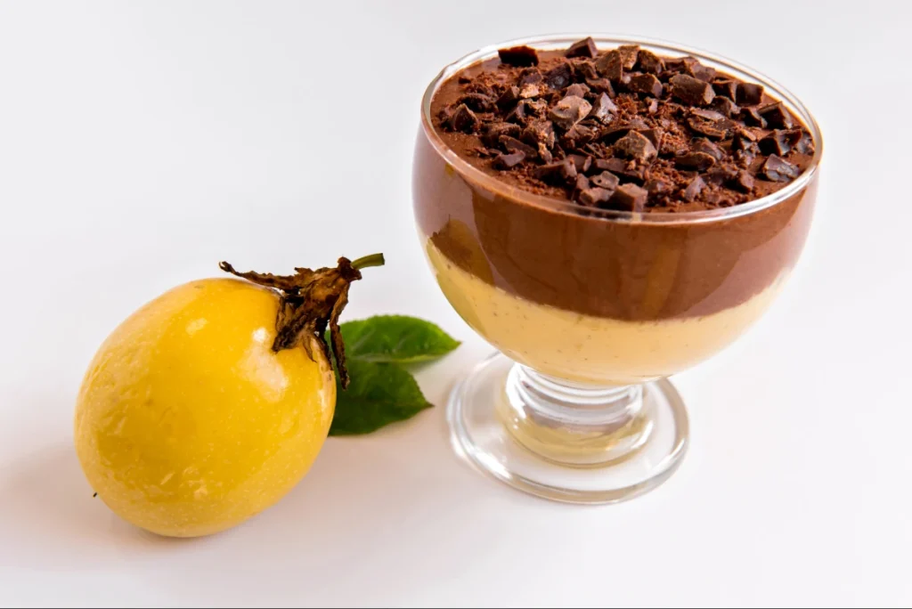 Sobremesa em camadas apresentando mousse de maracujá na base e uma densa camada de chocolate por cima, finalizada com chocolate picado. Um maracujá inteiro está ao lado.