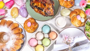 Mesa de almoço de Páscoa com bolo, pãe, salada, carne assada e ovos e flores compondo a decoração.