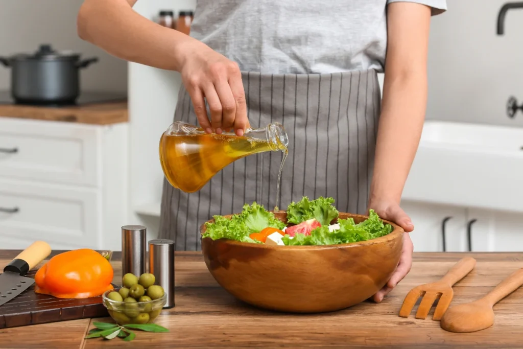 Mãos de uma pessoa usando avental despejam óleo de um frasco de vidro sobre uma salada de alface e tomates em uma tigela de madeira.