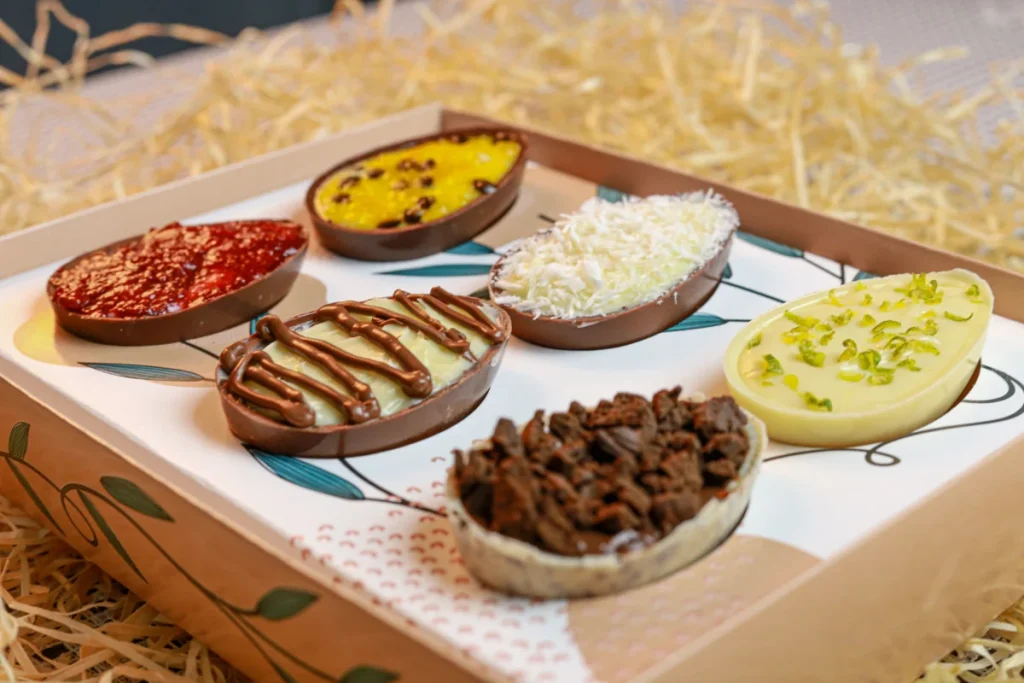 Vista superior de uma seleção de metades de ovos de chocolate recheados com sabores variados, como brigadeiro, maracujá, frutas vermelhas e confeitos crocantes, dentro de uma caixa.