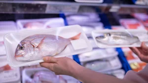 Home 2 essoa escolhendo peixe em supermercado. Os peixes estão em embalagens de isopor, cobertas por plástico filme, com características claras de peixe fresco.
