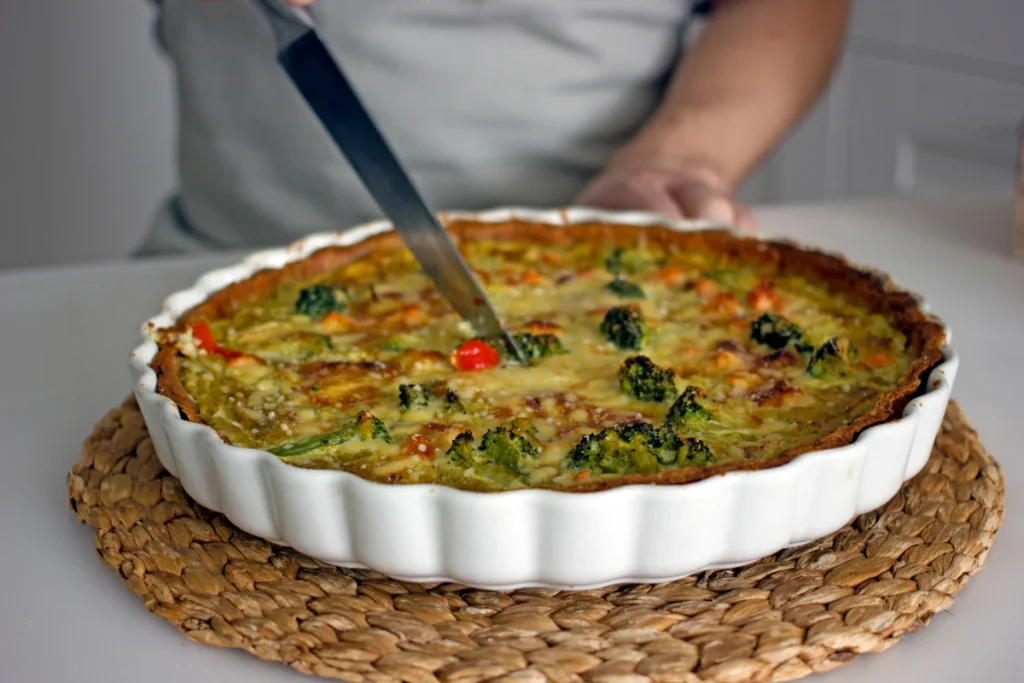  Quiche de brócolis e queijo gratinado sendo cortada em uma forma de cerâmica branca, uma receita de outono.