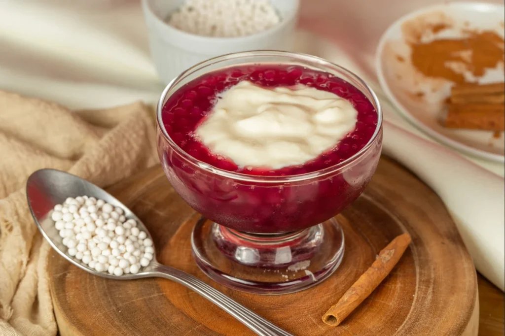 Taça de sagu servida ao lado de uma colher com bolinhas de sagu cruas (brancas) e canela.