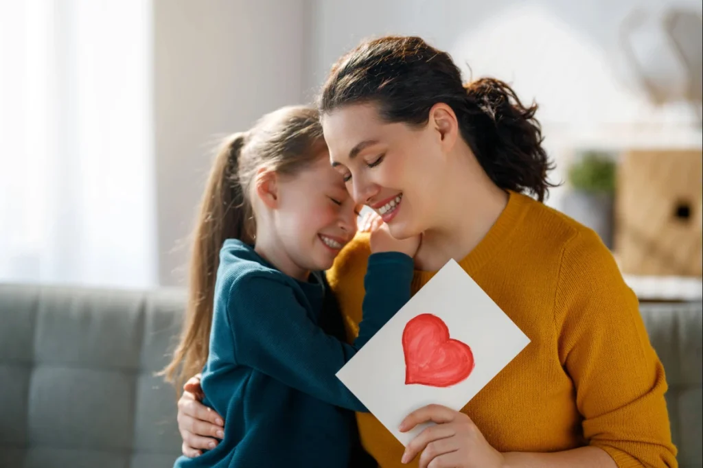 O que dar de presente no Dia das Mães? 24 ideias para todos os bolsos 3 Mãe e filha se abraçando enquanto a mãe segura um cartão com coração.
