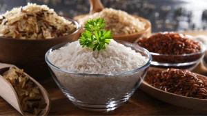 Tigela de vidro com arroz branco cozido decorado com salsa, cercada por outras variedades de arroz, como arroz integral, vermelho e selvagem em recipientes de madeira.