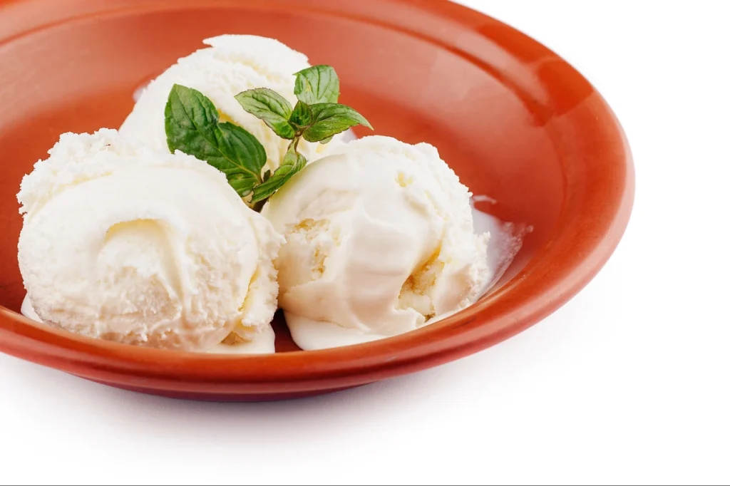 Três bolas de sorvete branco de creme ou baunilha servidas em um prato de cerâmica marrom, decoradas com folhas de hortelã fresca.