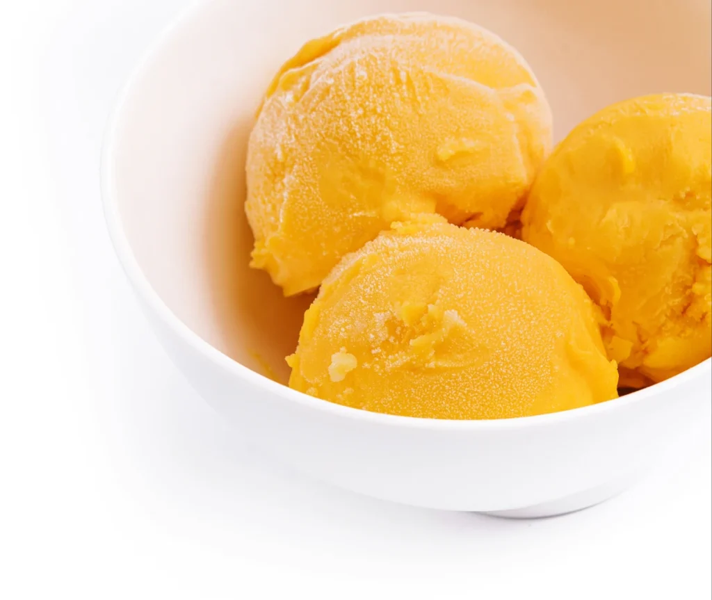 Tigela branca contendo três bolas de sorvete de manga amarelo, centralizadas sobre um fundo branco.