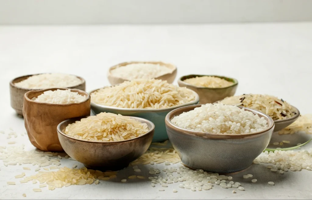 Diversas tigelas de cerâmica cheias com diferentes tipos de arroz cru, como arroz agulhinha, integral e parboilizado, espalhados sobre uma mesa clara.